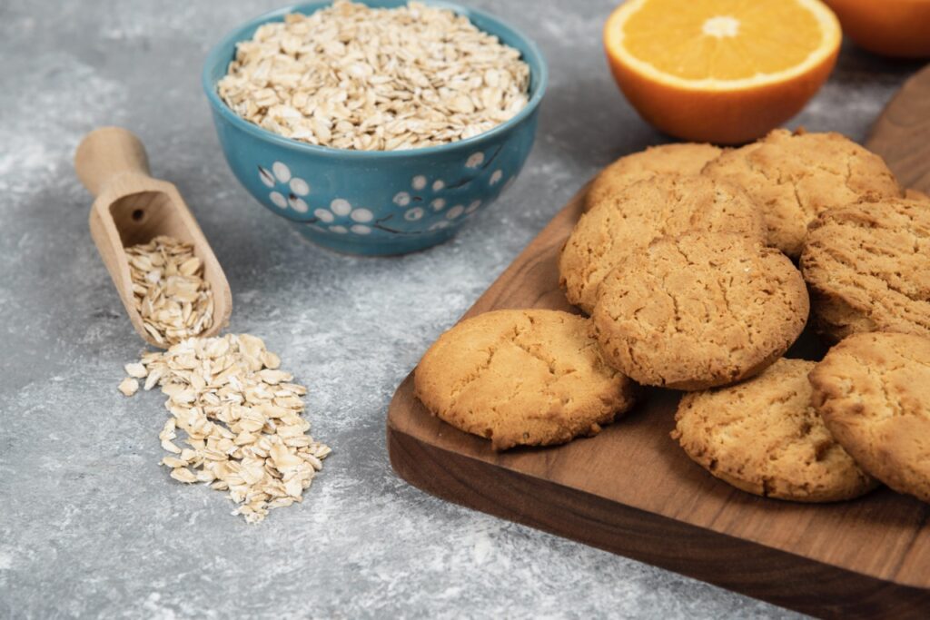 Biscoitos crocantes de aveia servidos em uma tábua de madeira, com aveia em flocos ao lado.