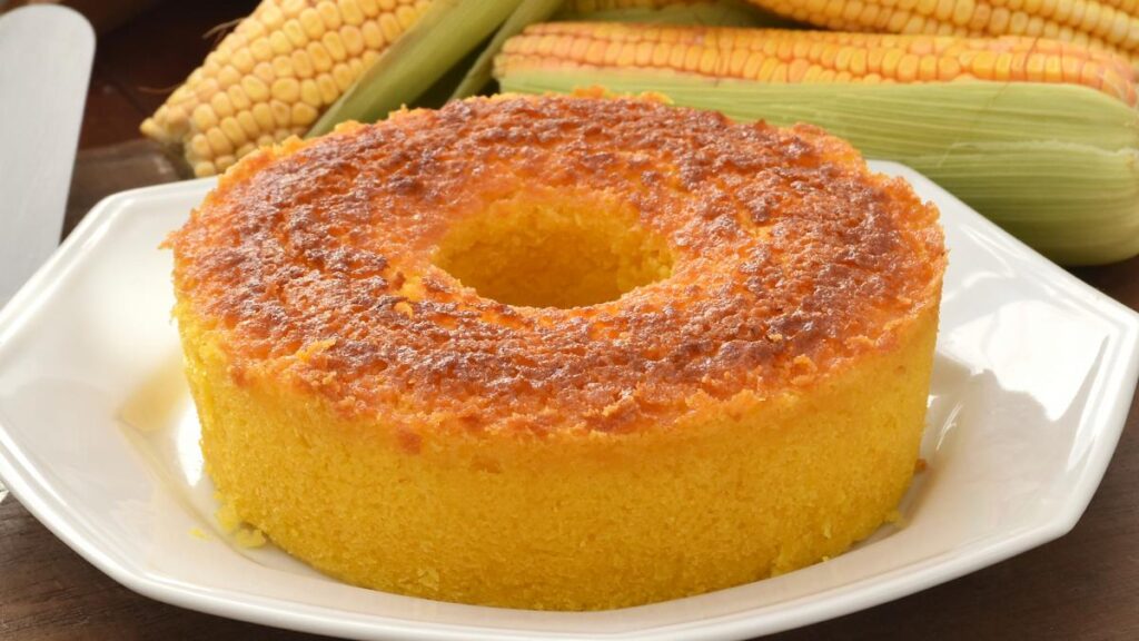 Bolo de milho cremoso em formato de anel, servido em prato branco com espigas de milho ao fundo.