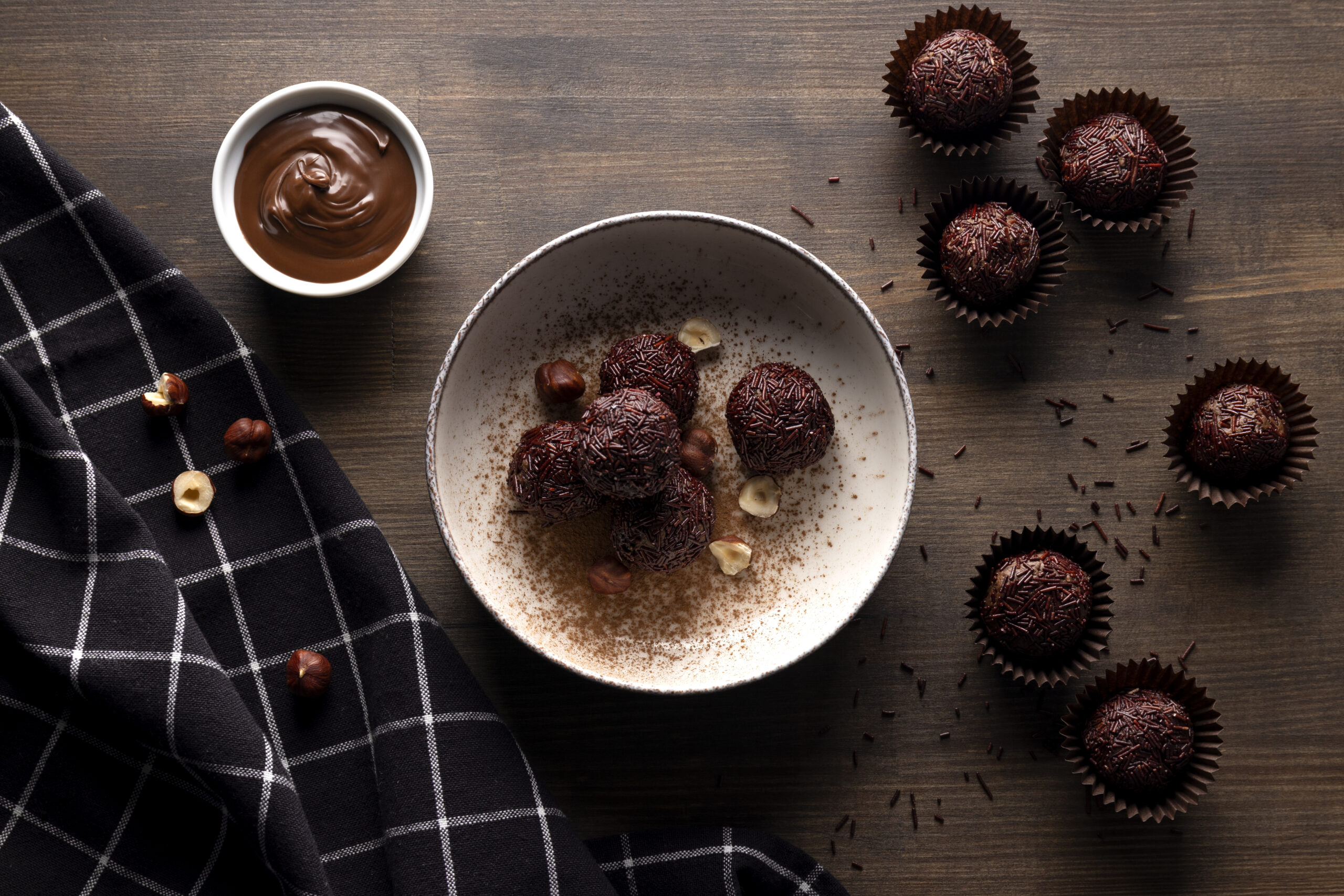 Brigadeiros low carb em uma tigela, decorados com granulado de cacau, ao lado de avelãs e um pote de chocolate derretido.