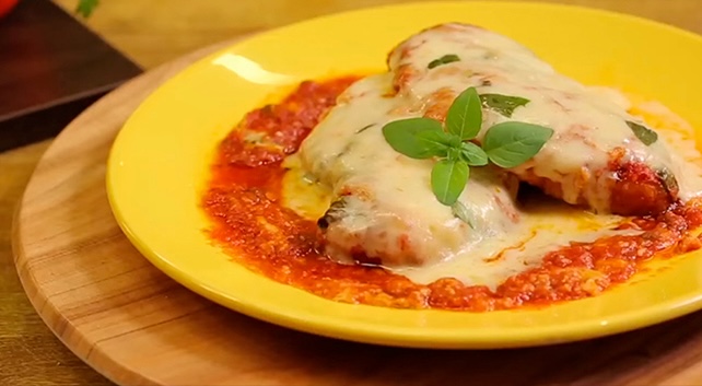Frango à parmegiana assado, coberto com queijo derretido e molho de tomate, servido em um prato amarelo com manjericão fresco como decoração.