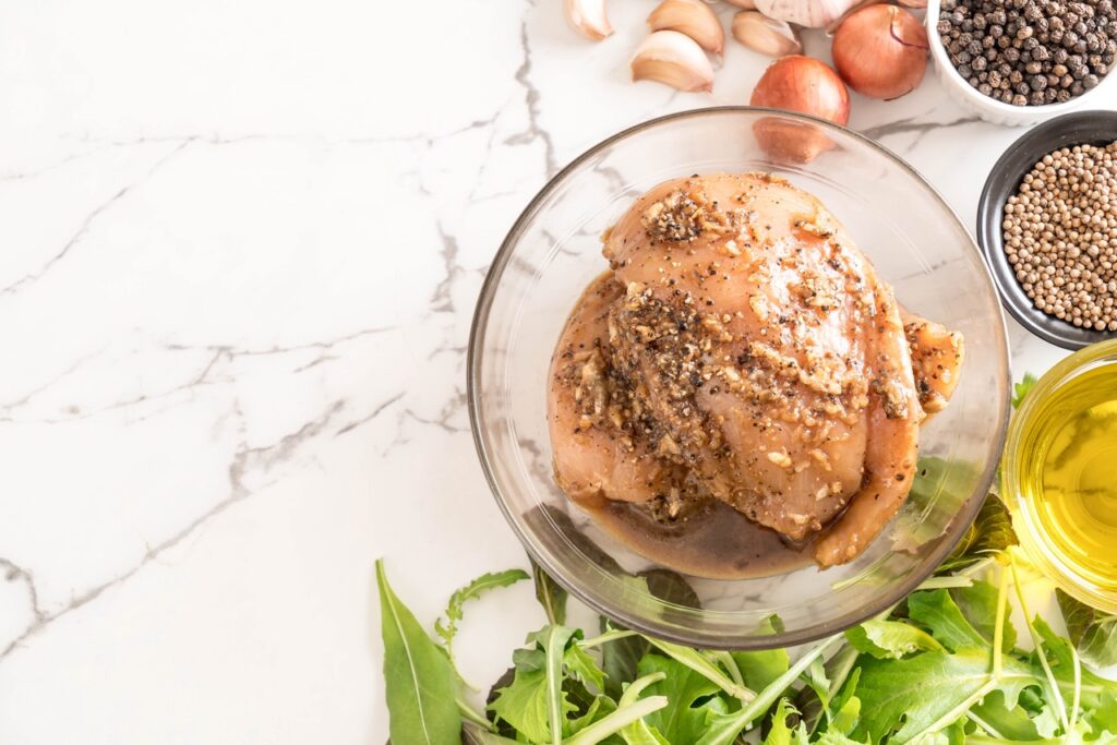Frango temperado com ervas finas e especiarias, preparado em uma tigela para marinada, acompanhado de alho, cebola e grãos de pimenta, ideal para uma refeição saudável e saborosa.

