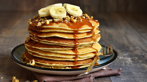 Panquecas empilhadas com cobertura de banana, nozes e calda de caramelo, servidas em um prato rústico.