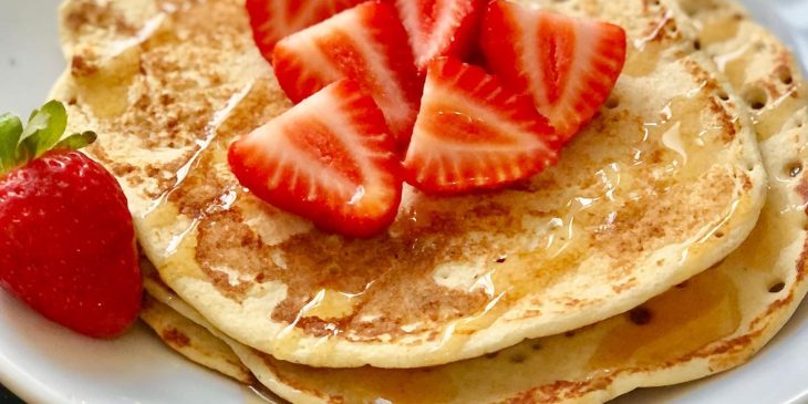 Panqueca proteica de whey coberta com fatias de morango e mel, servida em um prato branco.

