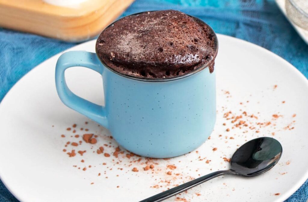 Bolo de caneca de chocolate servido em uma caneca azul, com uma colher ao lado.
