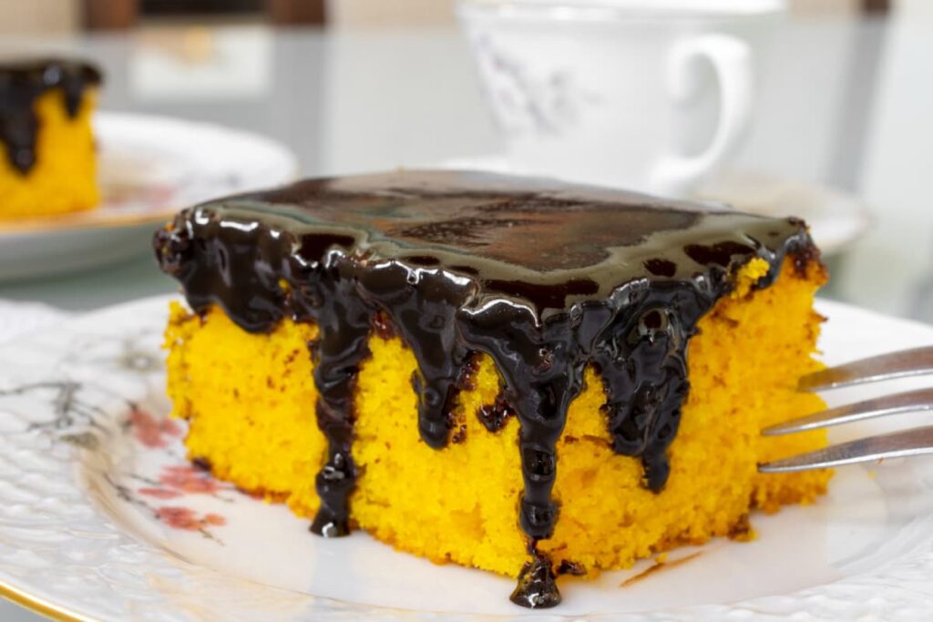 Fatia de bolo de cenoura com calda de chocolate escorrendo em prato decorado.

