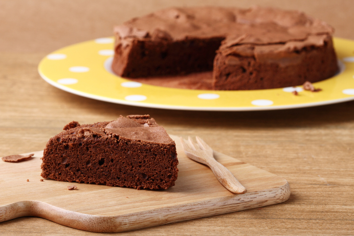 Fatia de bolo de chocolate proteico com whey servida em uma tábua de madeira, com o bolo inteiro ao fundo.