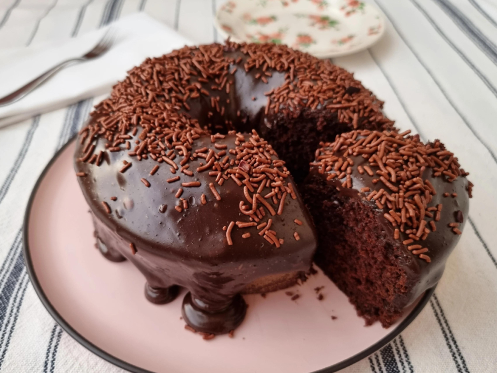 Bolo de chocolate com cobertura de brigadeiro e granulado, servido em prato rosa.