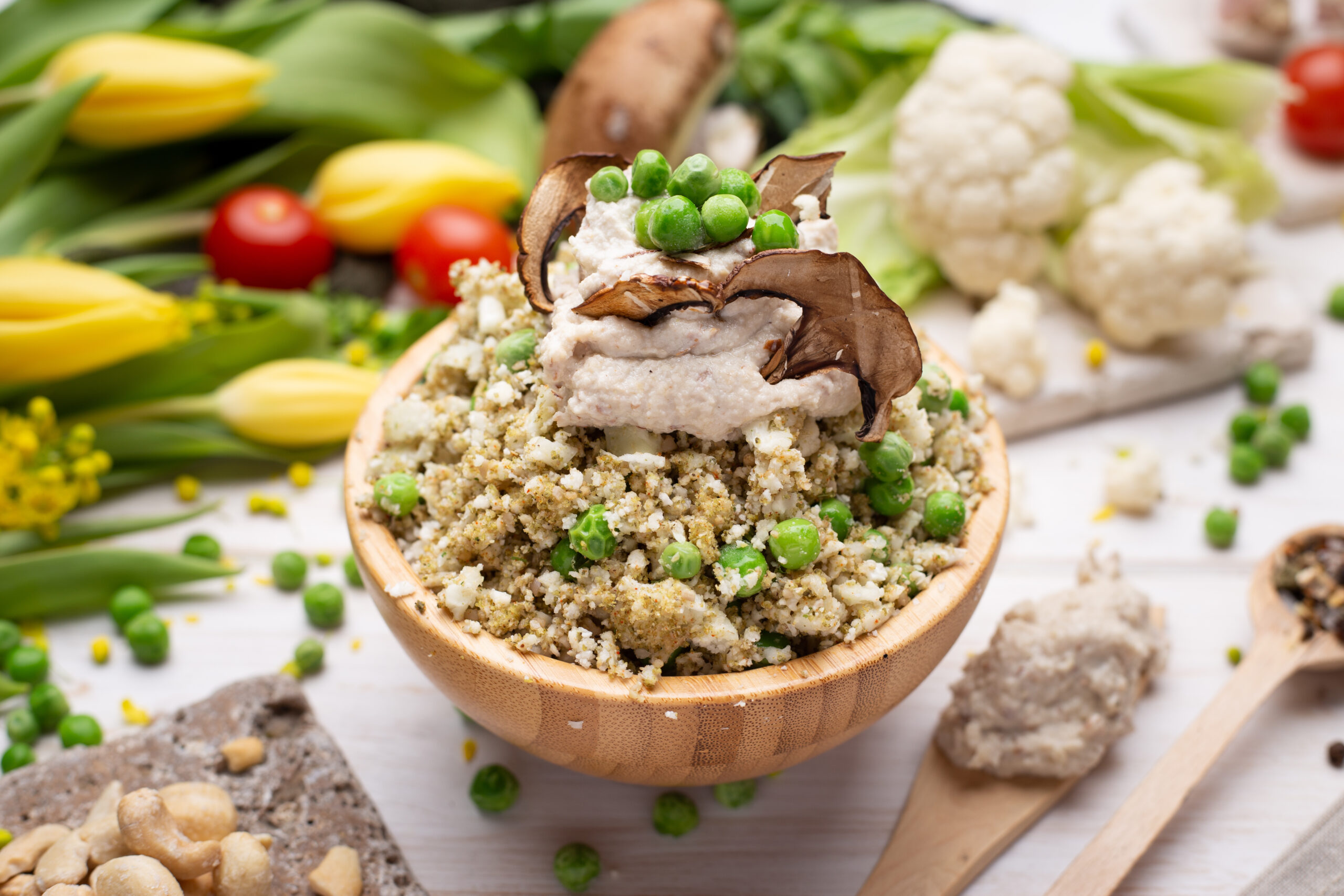 Arroz de couve-flor com cogumelos e ervilhas servido em uma tigela de madeira, decorado com um creme especial e cogumelos desidratados, em um ambiente decorativo com vegetais frescos.