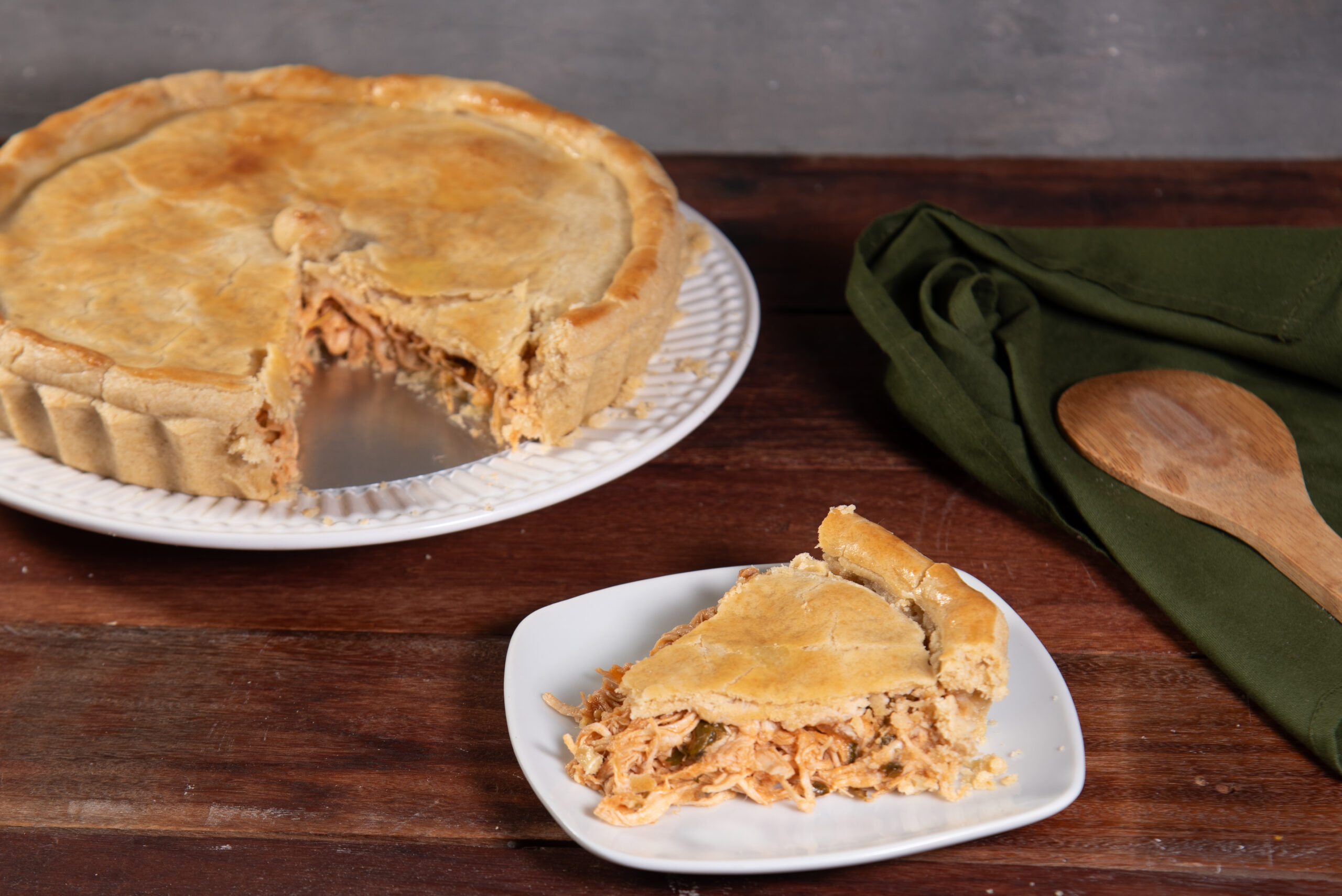 Empadão de frango cremoso com massa integral servido em um prato, com uma fatia destacada sobre um pires branco, em uma mesa rústica de madeira.
