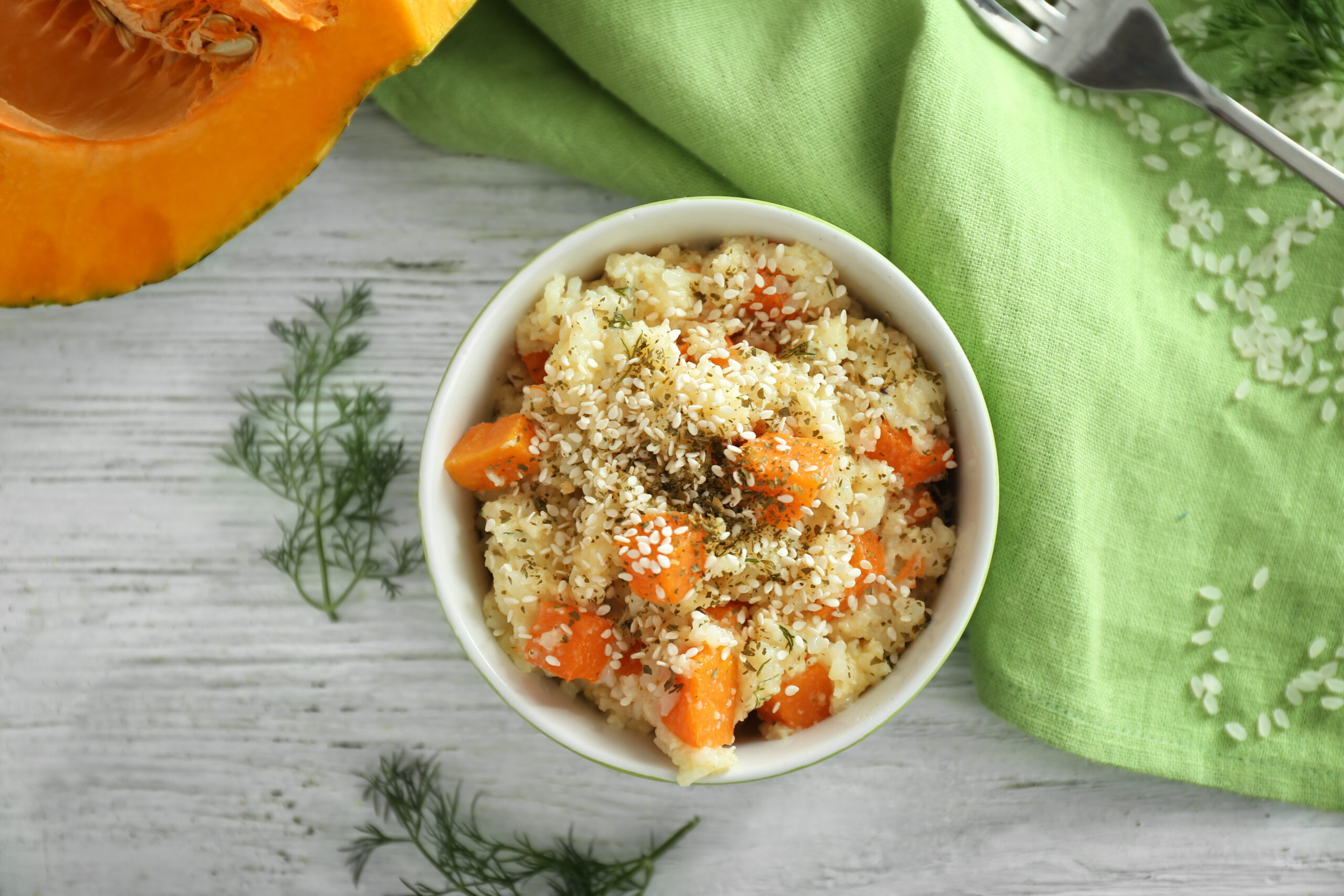 Prato de quinoa com legumes assados e frango desfiado, decorado com sementes e ervas frescas, servido em um bowl branco com um fundo de toalha verde e abóbora ao lado.