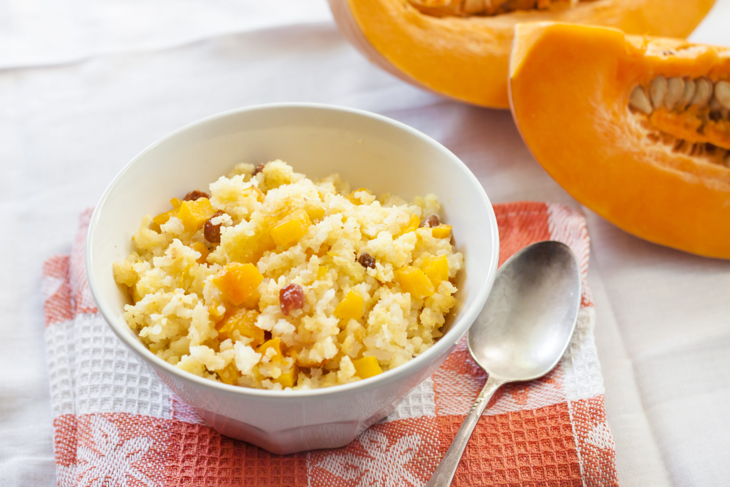 Prato de risoto de abóbora servido em uma tigela branca com pedaços de abóbora e passas, acompanhado de uma colher de metal e um pedaço de abóbora ao fundo.