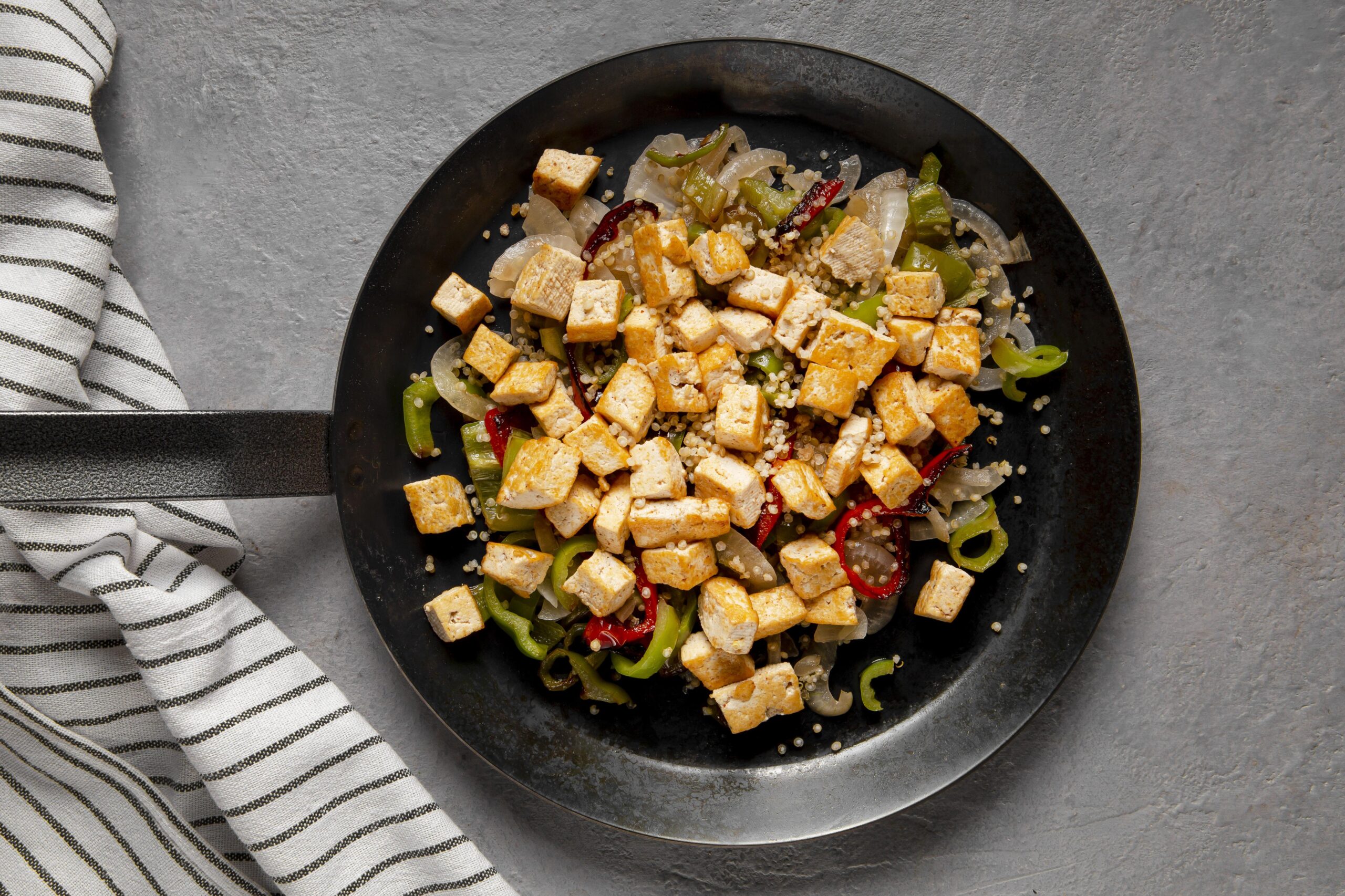 Tofu grelhado com legumes ao vapor servido em uma frigideira preta, acompanhado de pimentões, cebola e quinoa.