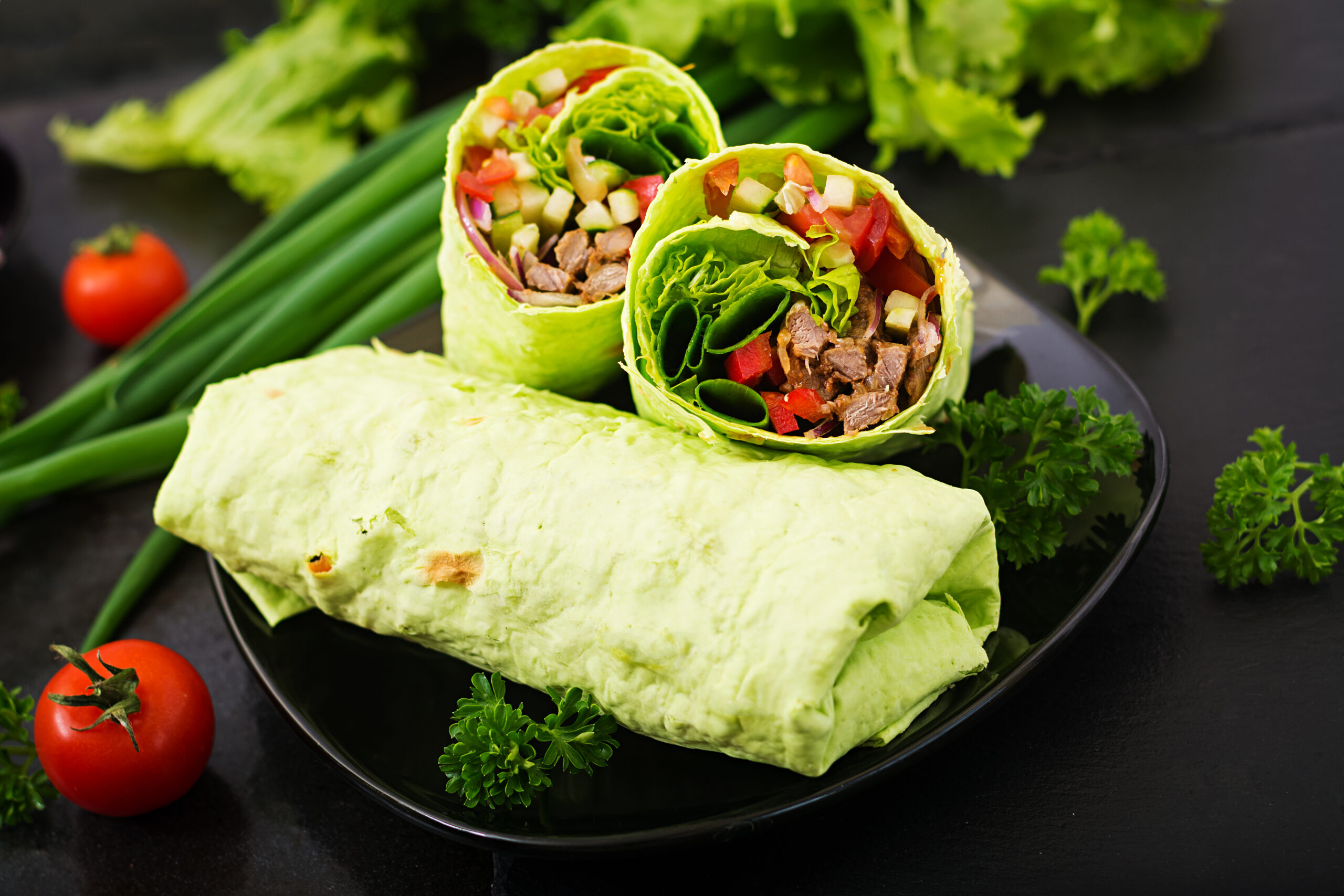 Wrap de atum recheado com folhas verdes, tomate e legumes, servido em um prato preto com guarnição de salsinha fresca.
