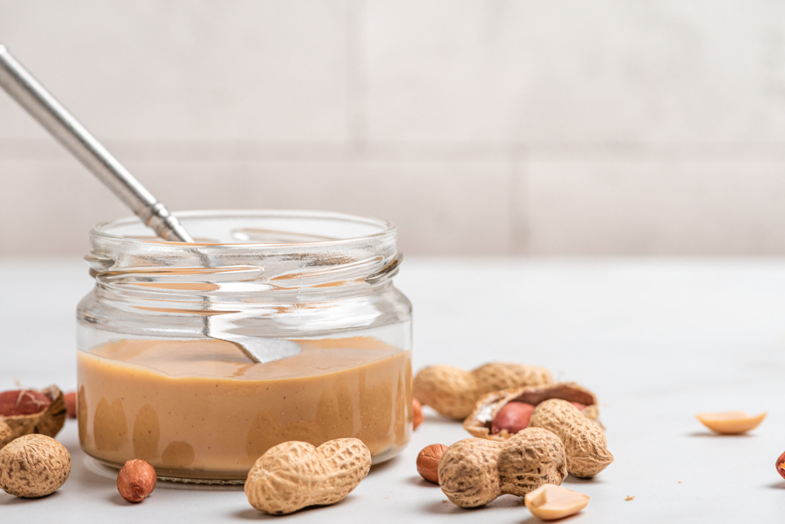 Pote de creme proteico de amendoim com whey acompanhado por amendoins inteiros sobre uma superfície clara.
