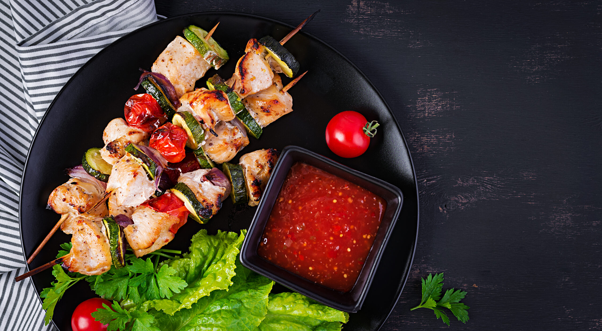 Espetinhos de frango com legumes grelhados, servidos com molho de tomate em um prato decorado com folhas verdes.