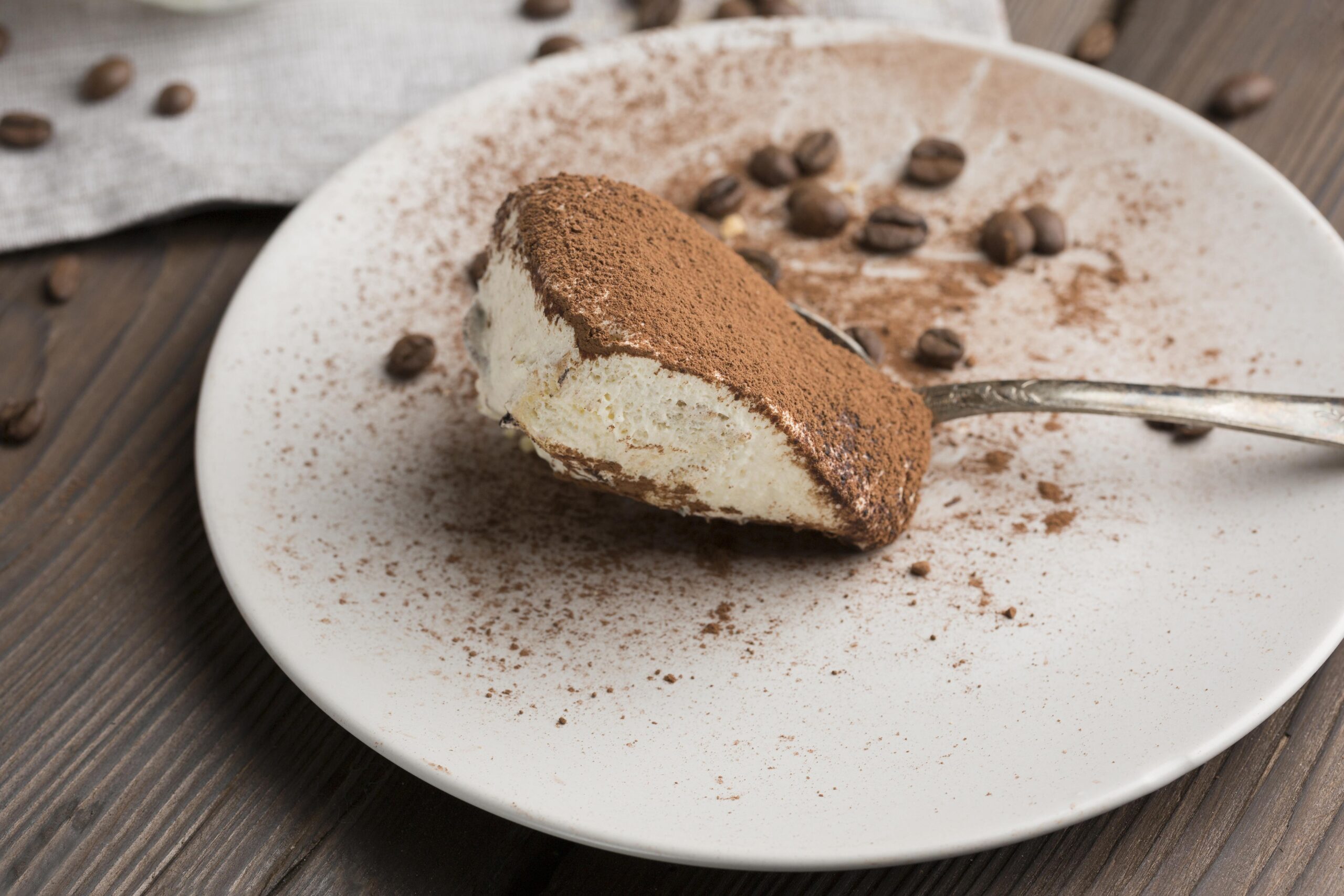 Fatia de tiramisù low carb com creme de mascarpone e cobertura de cacau em pó, servida em um prato com grãos de café ao redor.