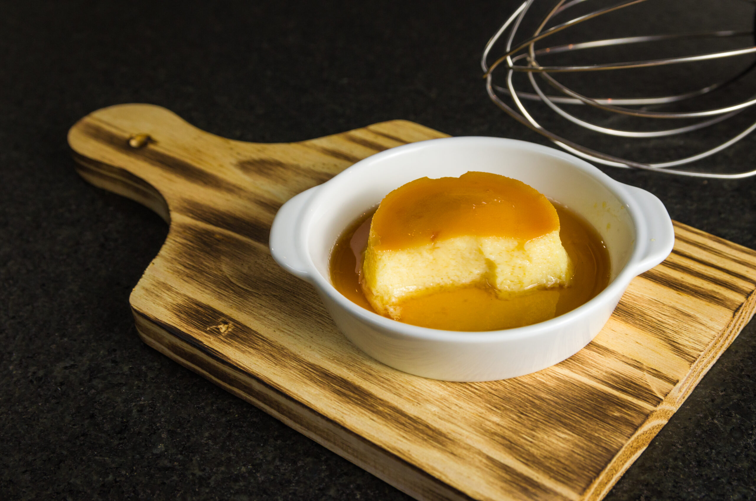Flan de baunilha sem açúcar servido em uma tigela branca com calda dourada sobre uma tábua de madeira.