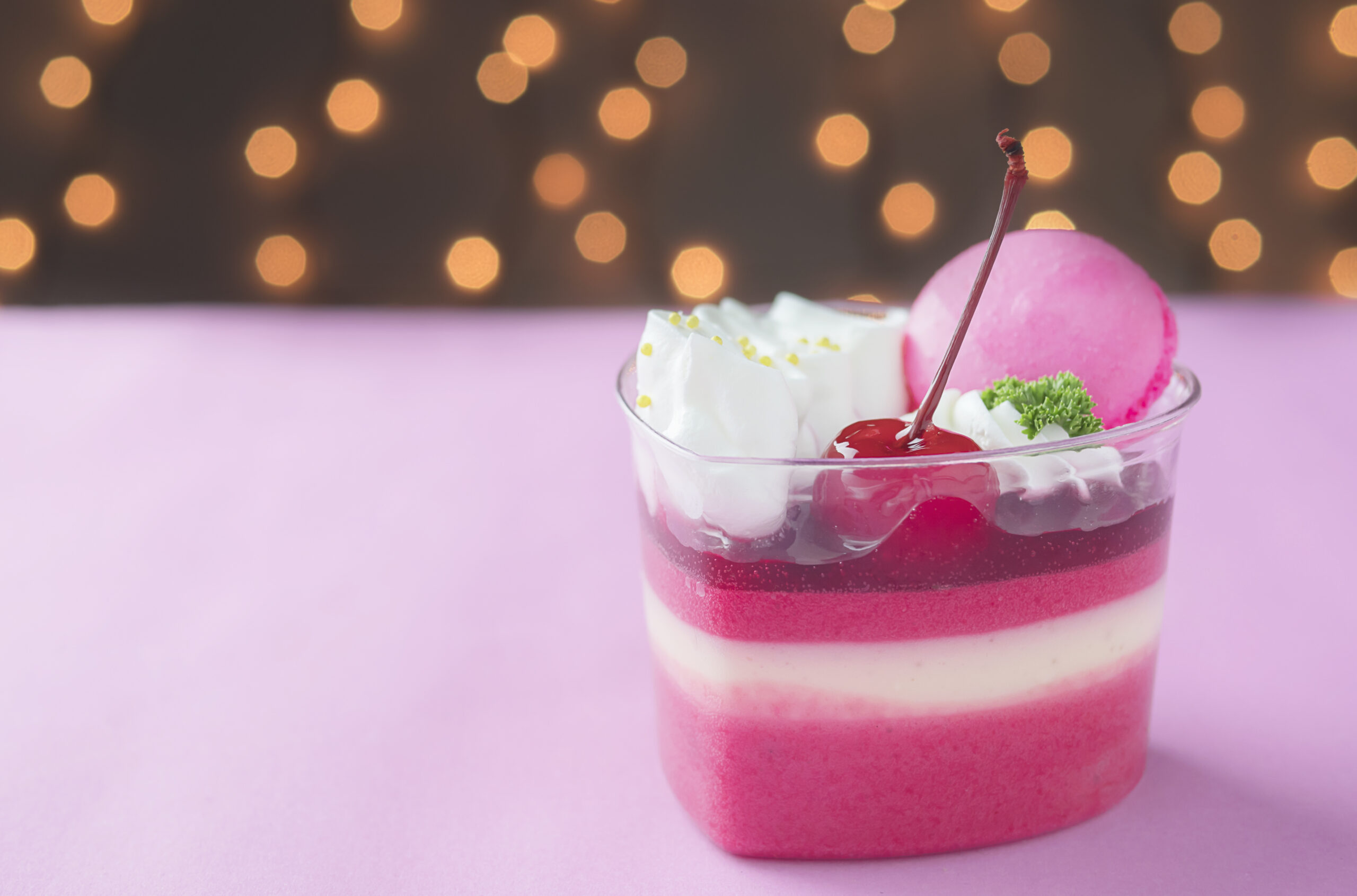 Taça de gelatina cremosa low carb com camadas de creme de ricota e decoração com chantilly, cereja, macaron rosa e enfeites dourados, sobre fundo rosa.