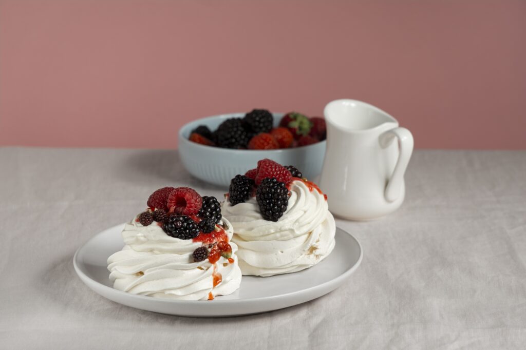 Pavlova de frutas vermelhas sem açúcar servida com um recipiente de creme e frutas frescas em destaque.
