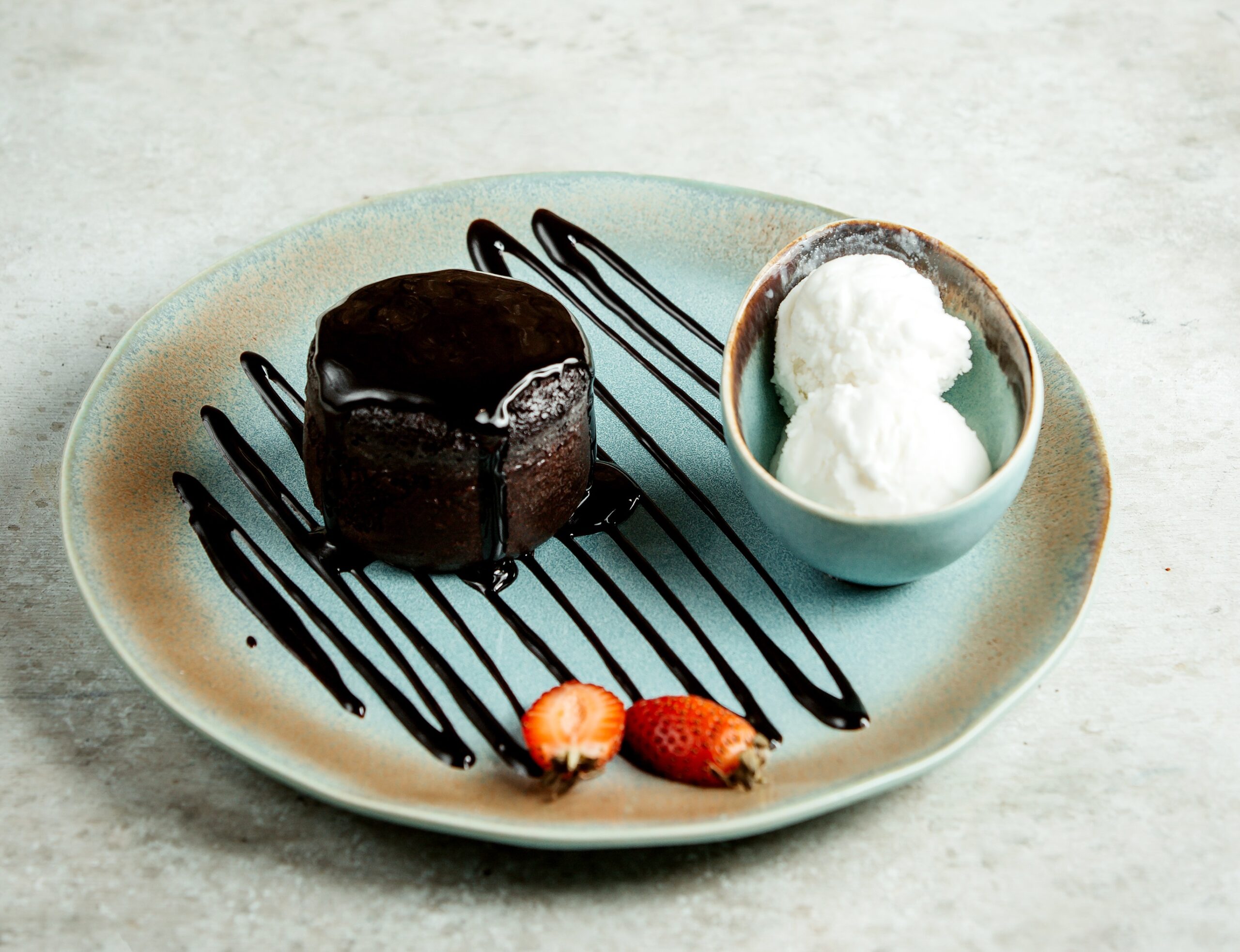 Petit gâteau low carb com cobertura de chocolate, acompanhado de sorvete de coco e morangos, servido em um prato azul claro com decoração de calda.